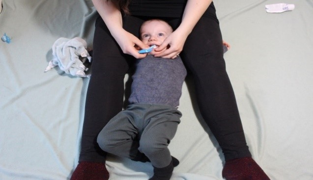 Det tredje billede viser en voksen siddende på gulvet med benene ud til siden. Barnet ligger med hovedet ind mod moderens mave og kigger op. Barnets arme er placeret under moderens ben. Barnets ben ligger frit. Moderen har et godt udsyn i barnets mund og børster barnets tænder i undermunden.